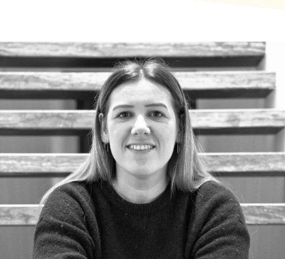 Vrouw met lang haar en oorbellen zit op een bank met houten banken op de achtergrond.