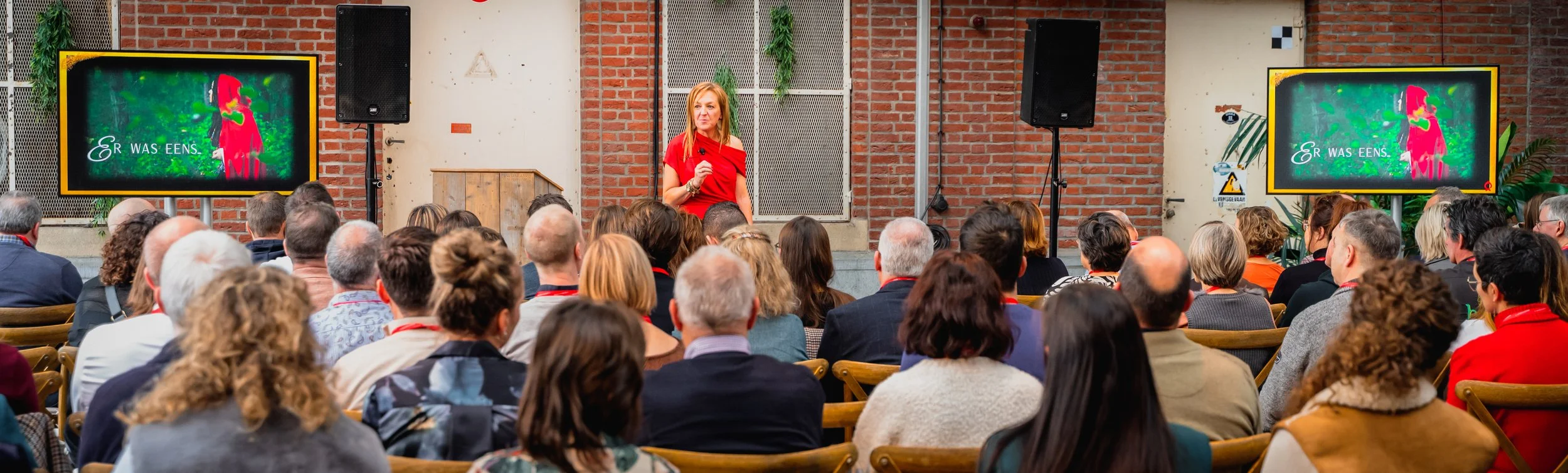Een vrouw geeft een presentatie aan een publiek in een zaal, met twee schermen op de achtergrond die een afbeelding en tekst tonen.