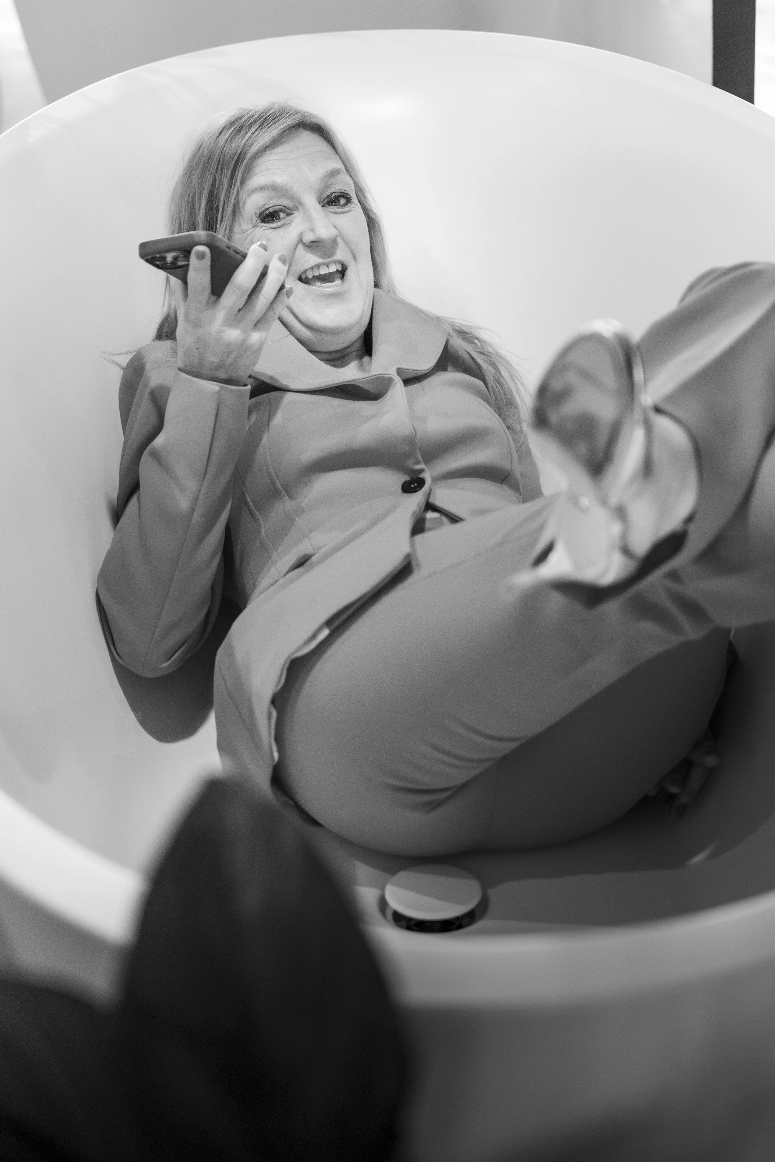 Vrouw ligt ontspannen in een bad, glimlacht en kijkt naar de camera, terwijl ze haar telefoon vasthoudt.