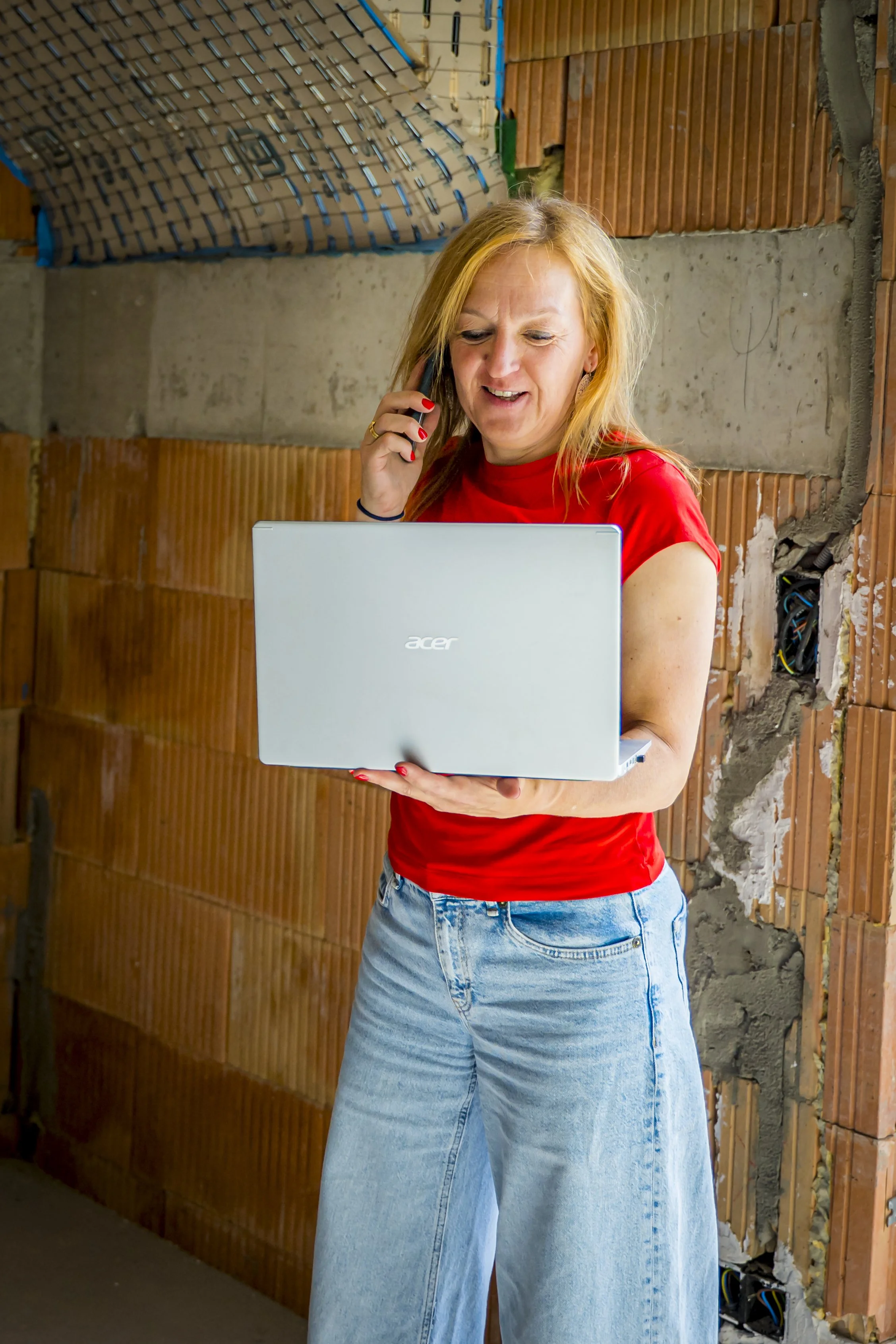Vrouw in rode t-shirt en blauwe jeans houdt een laptop vast en spreekt op de telefoon, in een bouwplaats of ruw afgewerkt interieur met bakstenen en betonmuur.