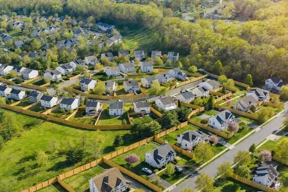 Aerial view of a suburban neighborhood with numerous single-family homes, well-maintained yards, and wooden fences, surrounded by green trees and wooded areas.