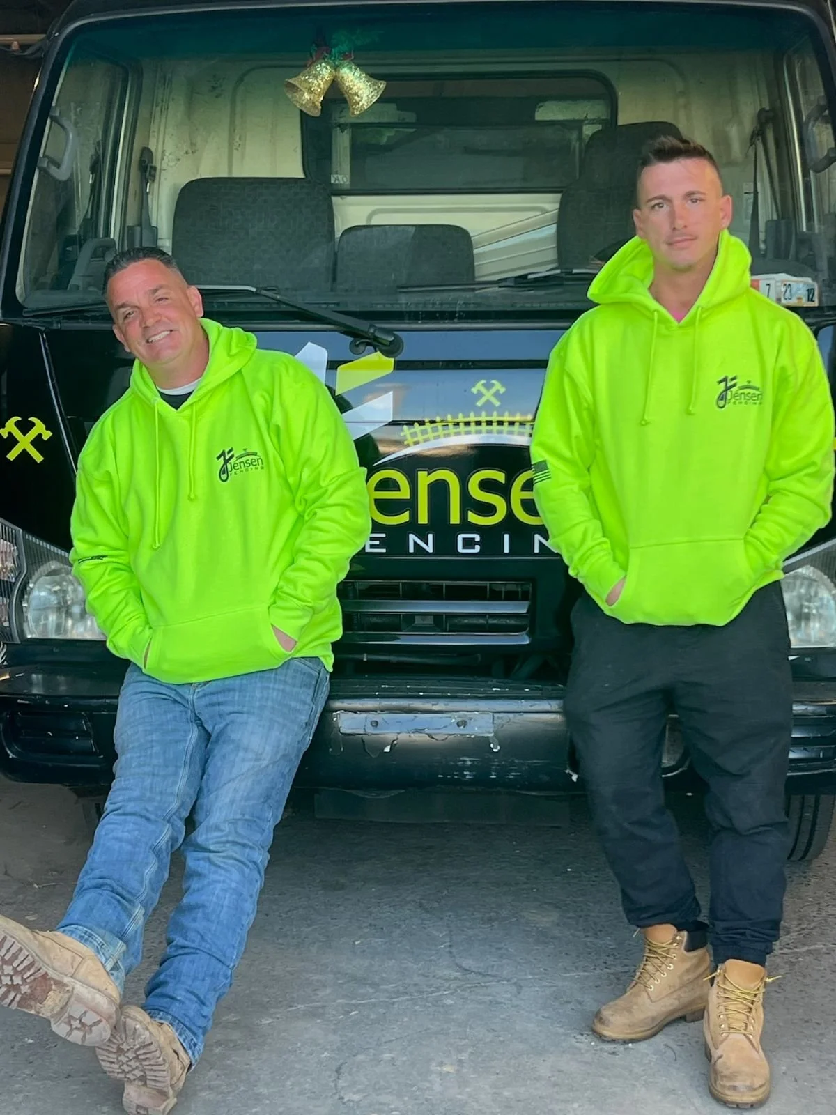 Two men in bright yellow hoodies and work boots leaning against a black vehicle with branding and crossed tools logo, standing outside on a dirt surface.