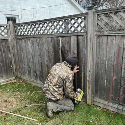 A person kneeling on the grass, using a power drill on a wooden fence in a backyard.