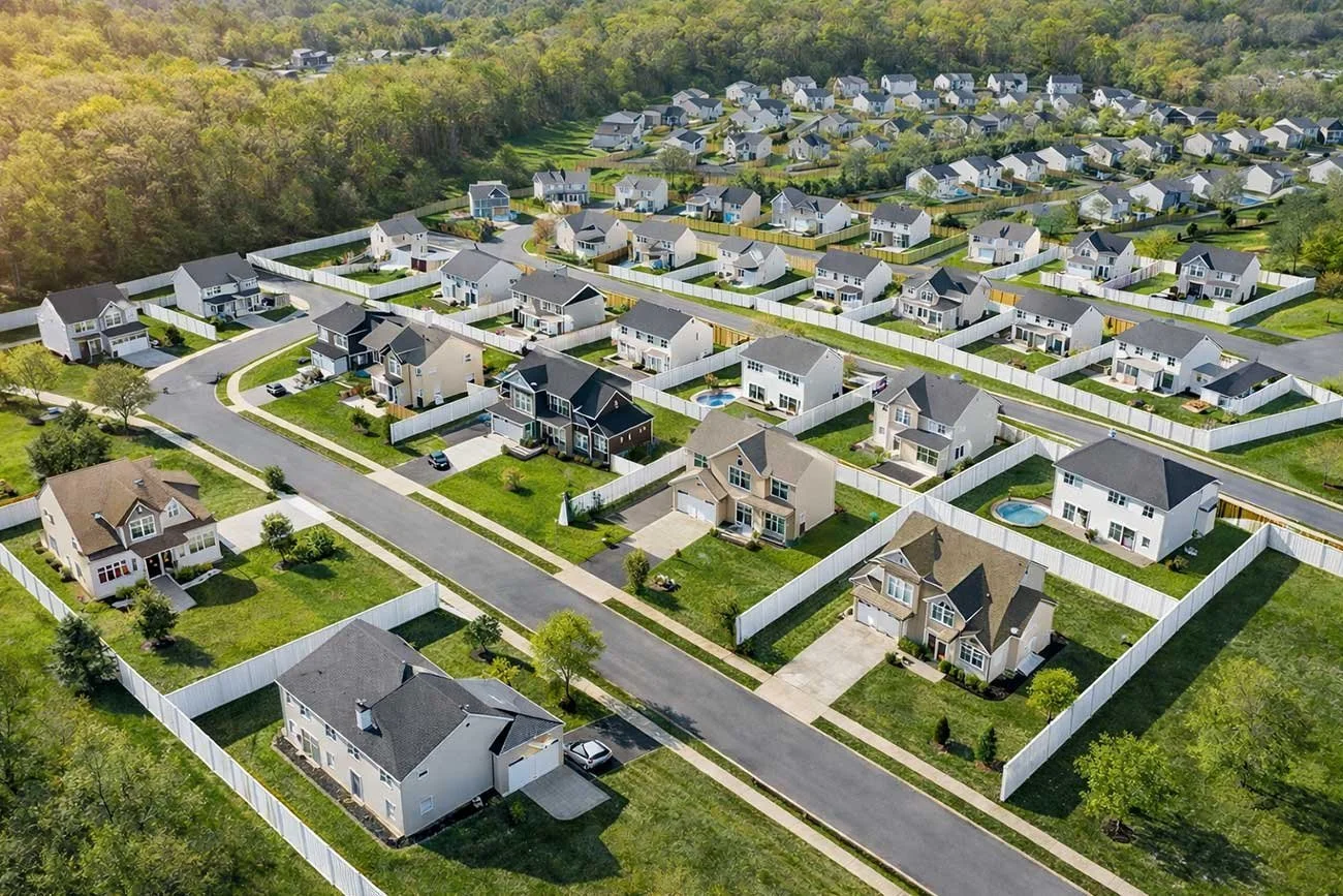 Aerial view of a suburban neighborhood with single-family homes, green lawns, fences, and streets, surrounded by trees.