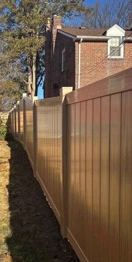 View of a wooden privacy fence running alongside a house with a brick exterior, under a clear blue sky, with trees in the background.