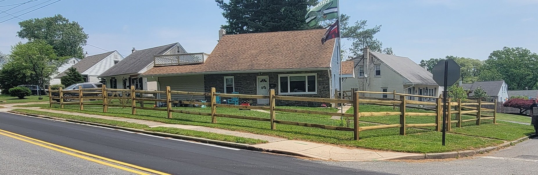 A residential house with a brown shingle roof, stone and siding exterior, surrounded by a wooden fence. The front yard has grass and sidewalk, with neighboring houses visible in the background. A stop sign and flags are near the house. Cars are parked along the street.