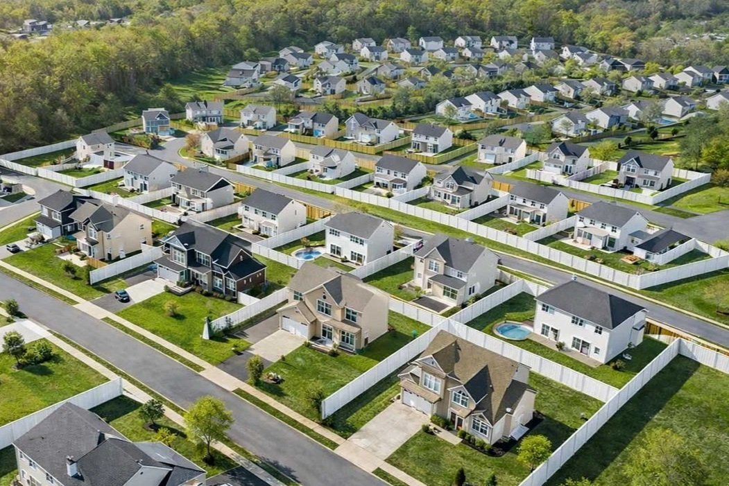 Aerial view of a suburban neighborhood with rows of single-family homes and fenced backyards, some with swimming pools, surrounded by trees.