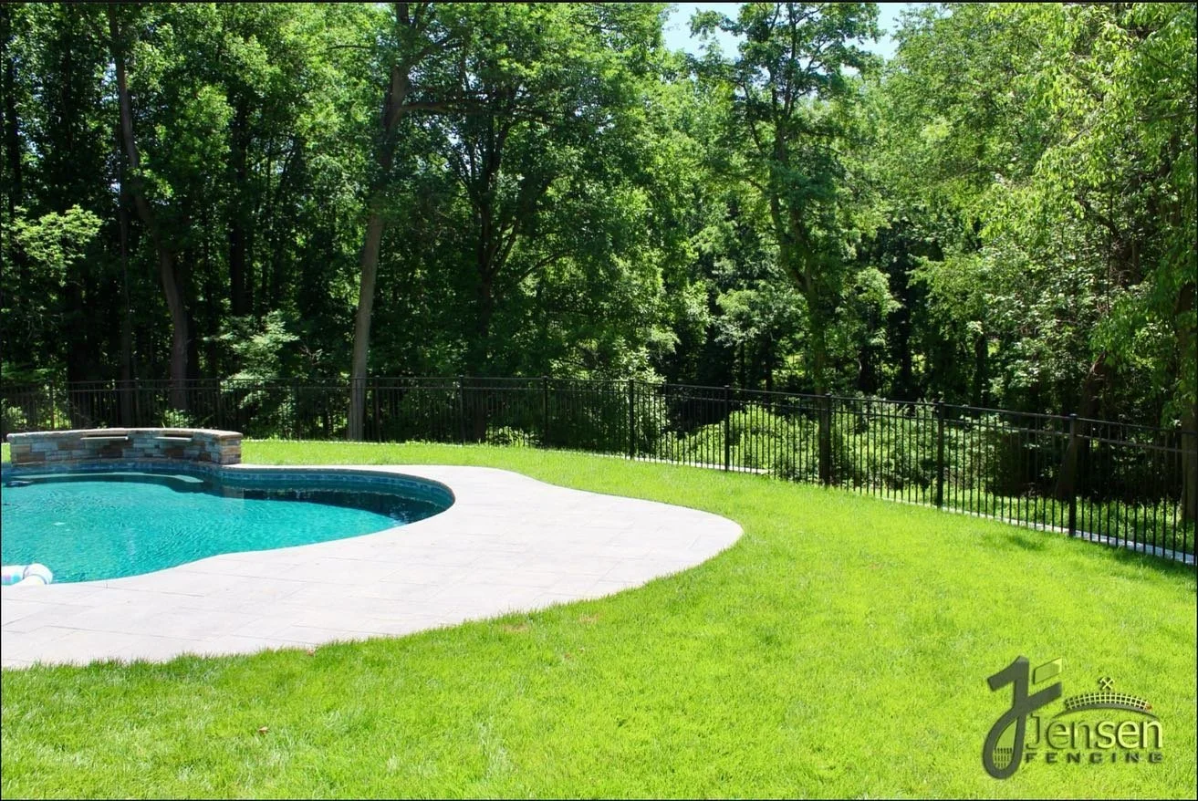 Backyard with a swimming pool, a stone waterfall feature, a grassy lawn, and a black metal fence, with tall trees in the background.