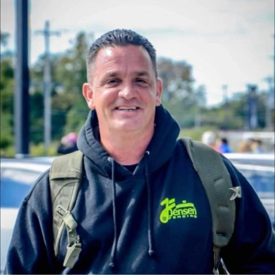 A smiling man in a black hoodie with a backpack outdoors, with trees and power lines in the background.