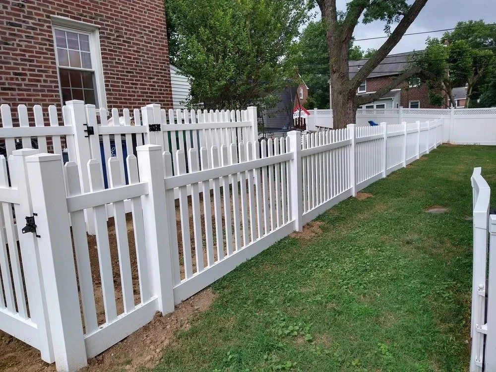 A white vinyl privacy fence surrounds a backyard with a brick house on the left and other houses and trees in the background. The yard has green grass and a small dirt patch near the fence.