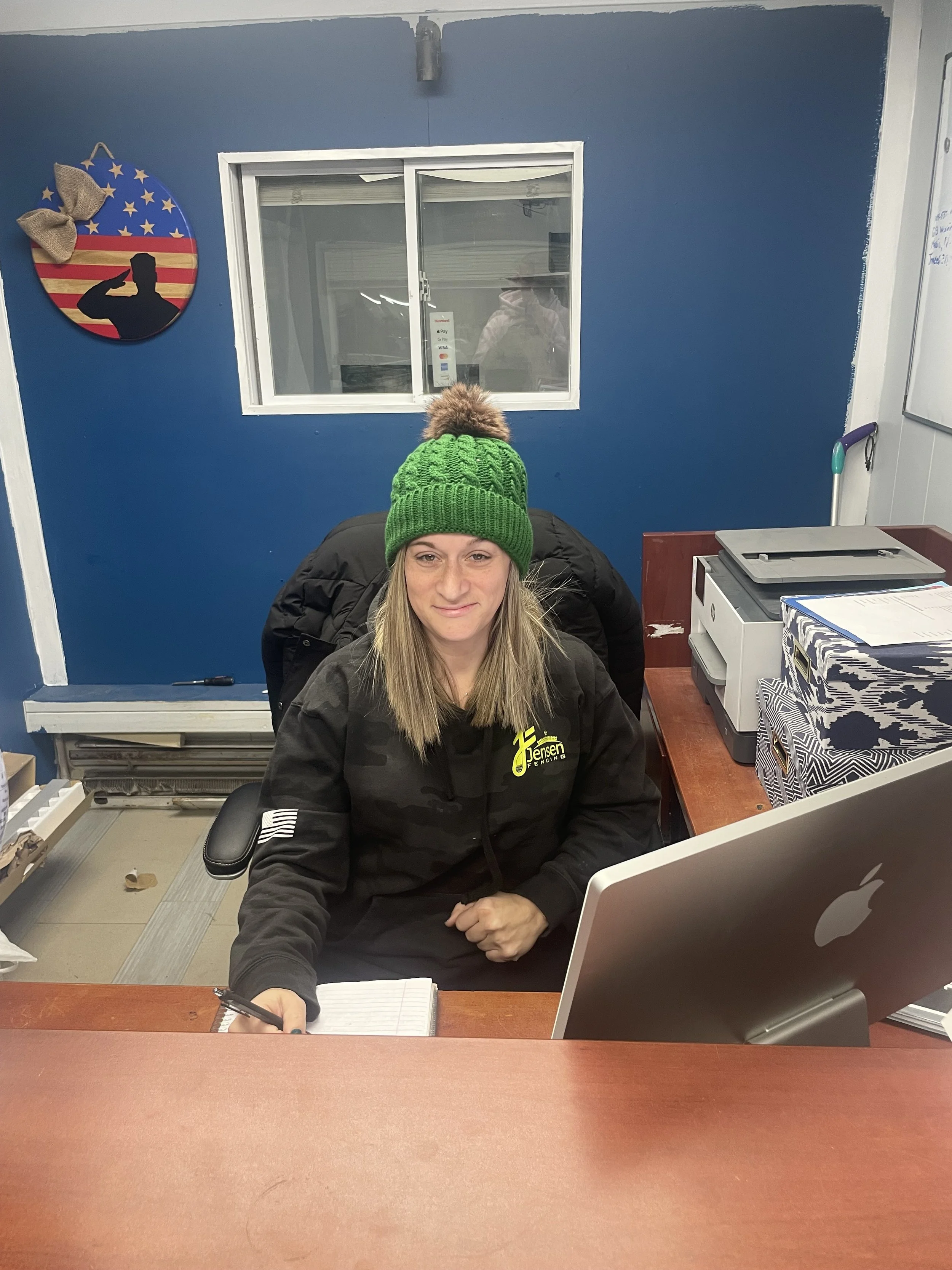 A woman sitting at a desk with a computer, wearing a green knit hat and a black jacket, in an indoor office with blue walls.
