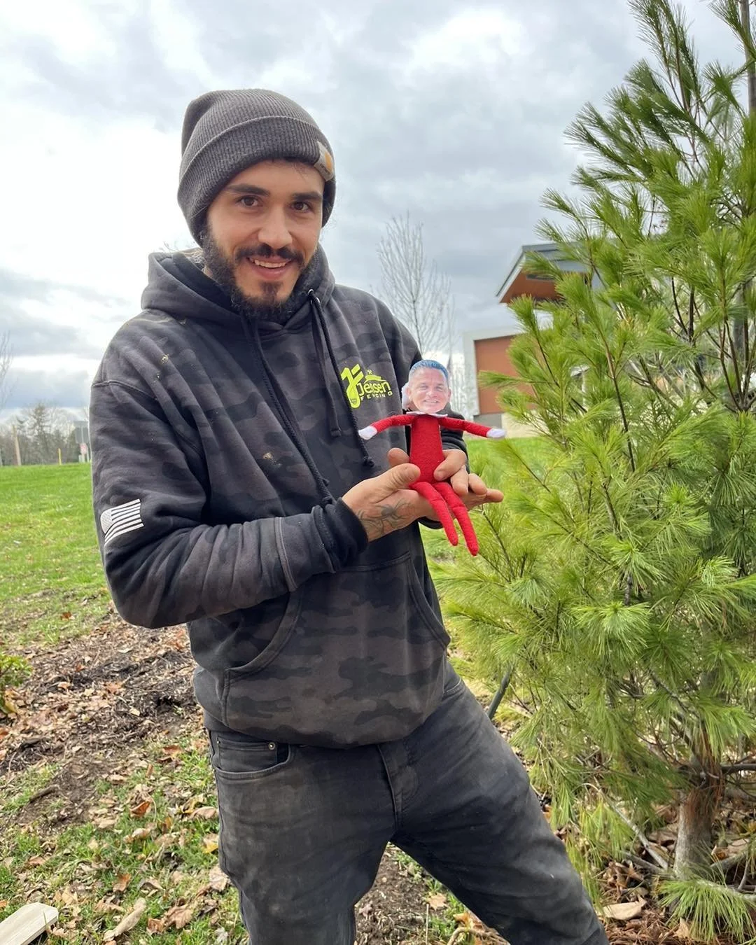A young man with a beard, wearing a gray beanie and black camouflage hoodie, stands outdoors next to a green shrub. He is holding a small doll with a photograph of a man's face on its head, dressed in red. The man is smiling and looking at the camera.