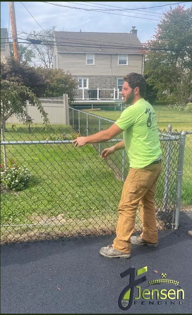 A man in tan pants and a bright yellow T-shirt stands outdoors, holding a metal fence with his right hand, looking towards his right side. The background includes a house with gray siding, a white fence, and some trees. The ground is paved asphalt, with a flower bed on the left and a logo in the bottom right corner that says 'Jensen Fences'.