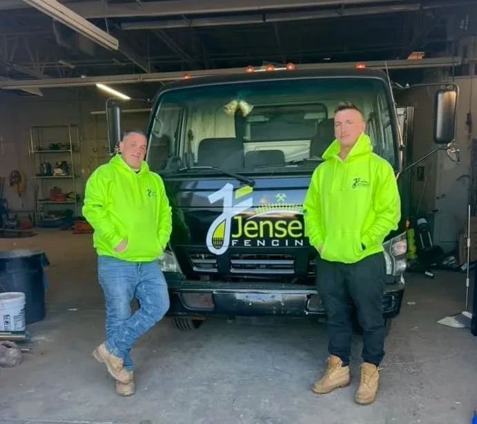 Two men in bright green hoodies standing in front of a black work truck inside a garage, with shelves and tools in the background.
