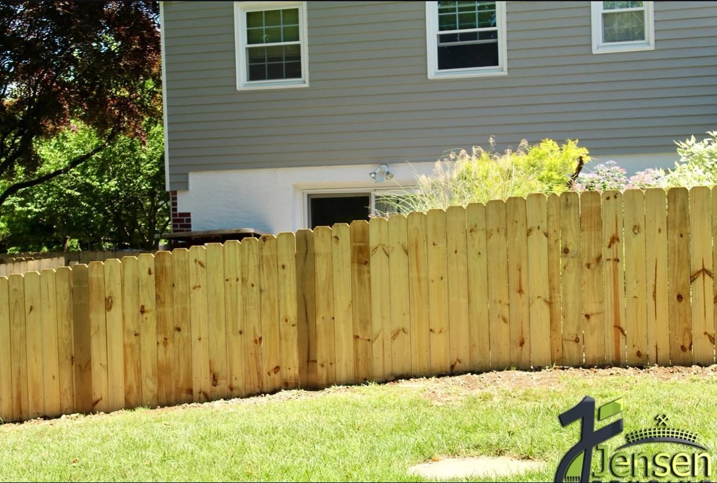 A backyard with a wooden fence, a house wall with three windows, and greenery including trees and plants.