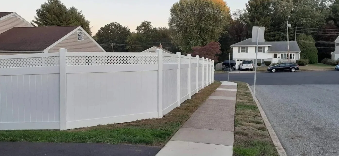 A sidewalk running alongside a white vinyl privacy fence in a residential neighborhood, with houses, trees, and parked cars in the background.
