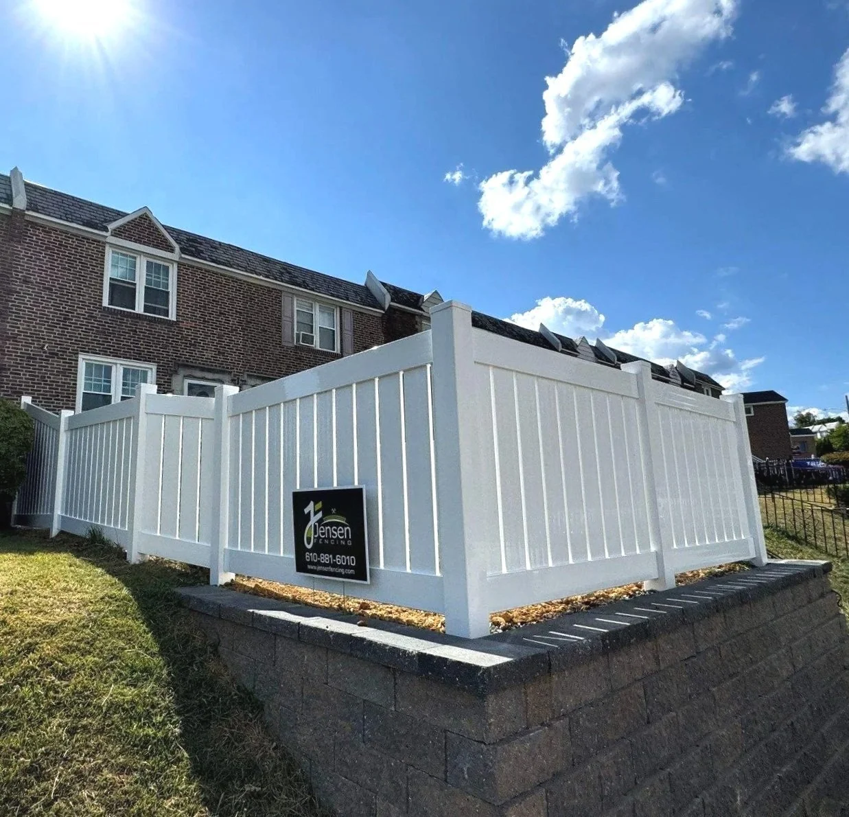 A white privacy fence surrounding a yard with a brick house in the background. The fence is installed on top of a raised stone foundation. A sign on the fence displays the company name 'Jensen Fencing' and contact information.