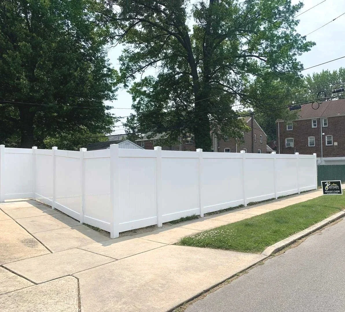 A white vinyl privacy fence surrounding a corner lot with a sidewalk and grassy area. There is a sign in the yard indicating a fencing company. Large trees and residential buildings are visible in the background.