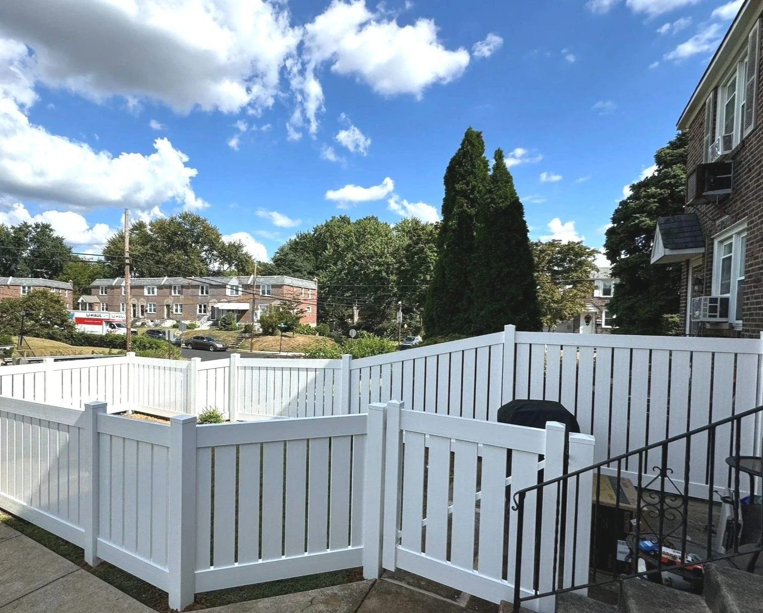 View of a residential backyard enclosed by a white picket fence, with a barbecue grill and steps leading down from a porch, under a partly cloudy sky with large trees and neighboring houses in the background.