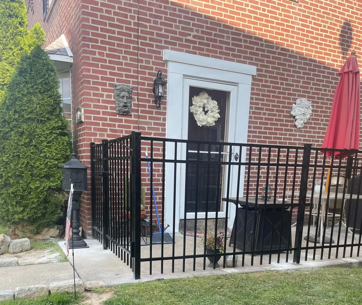 A small fenced patio in front of a brick house with white door frame and floral wreath, decorative lion heads on brick wall, a lantern, a black table, and a red umbrella.