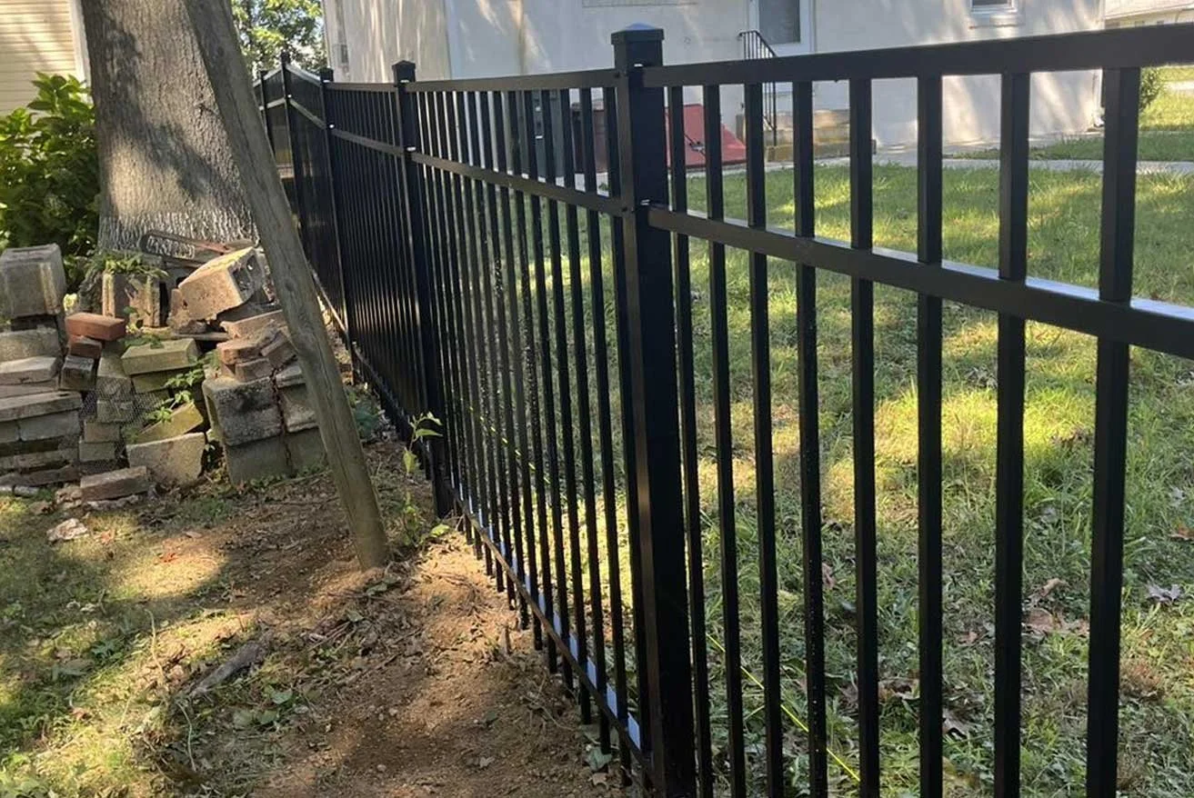 A black metal fence running along a grassy yard, with a tree and pile of bricks on one side.