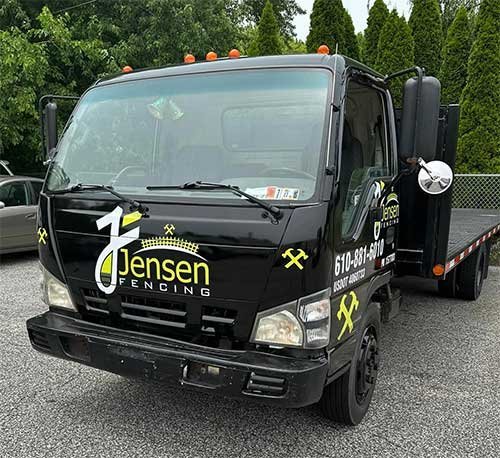 A black Jensen Fencing service truck parked on a gravel surface with trees in the background. The truck has a flatbed and the Jensen Fencing logo and contact information displayed on the door and hood.