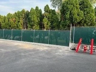 A fenced outdoor area with green mesh fencing, red traffic cones, and a paved surface, surrounded by tall trees.