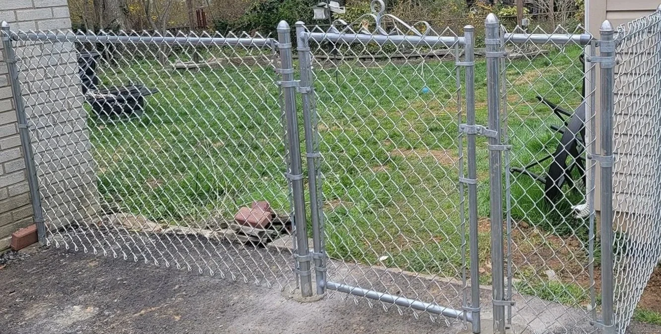 A chain-link fence with a small gate, enclosing a backyard with grass. On the left side next to the fence is a black trash can, and inside the fenced area on the right side are two dogs, one black and white and the other mostly black, standing near the grass.