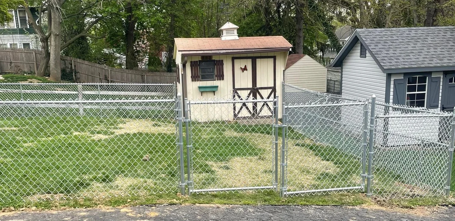 A backyard with a small beige shed featuring dark brown shuttered windows, a butterfly decoration, and a double gate, surrounded by a chain-link fence and neighboring houses with trees in the background.