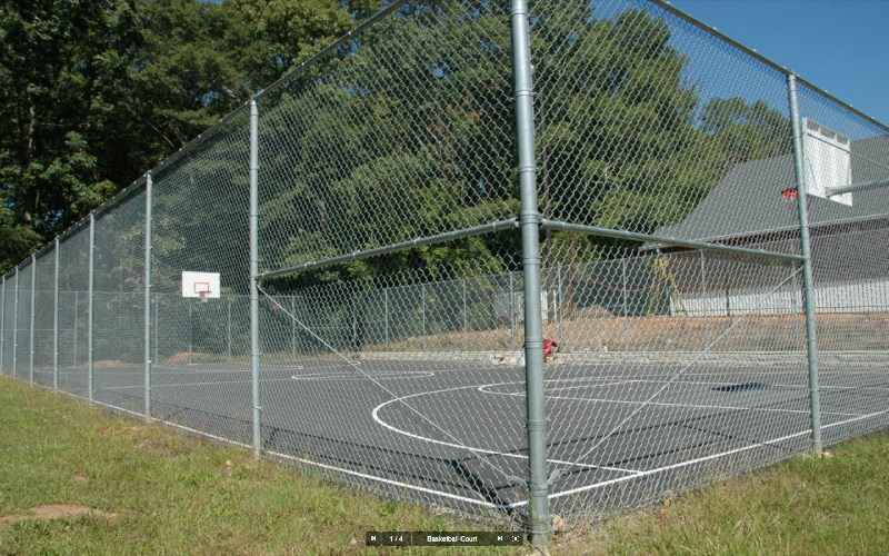 An outdoor basketball court surrounded by a chain-link fence with a basketball hoop in the background, trees, and a building.