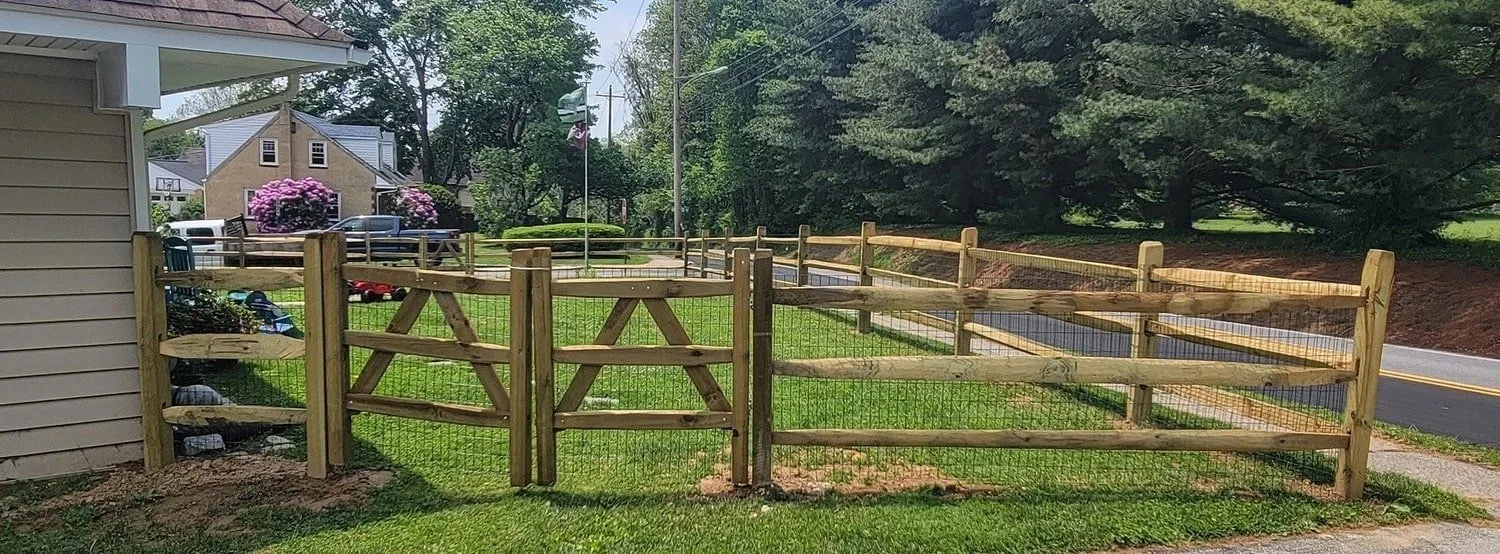 Wooden and wire mesh backyard fence with a gate, next to a house, with a lawn and a sidewalk, on a sunny day with trees and street in the background.