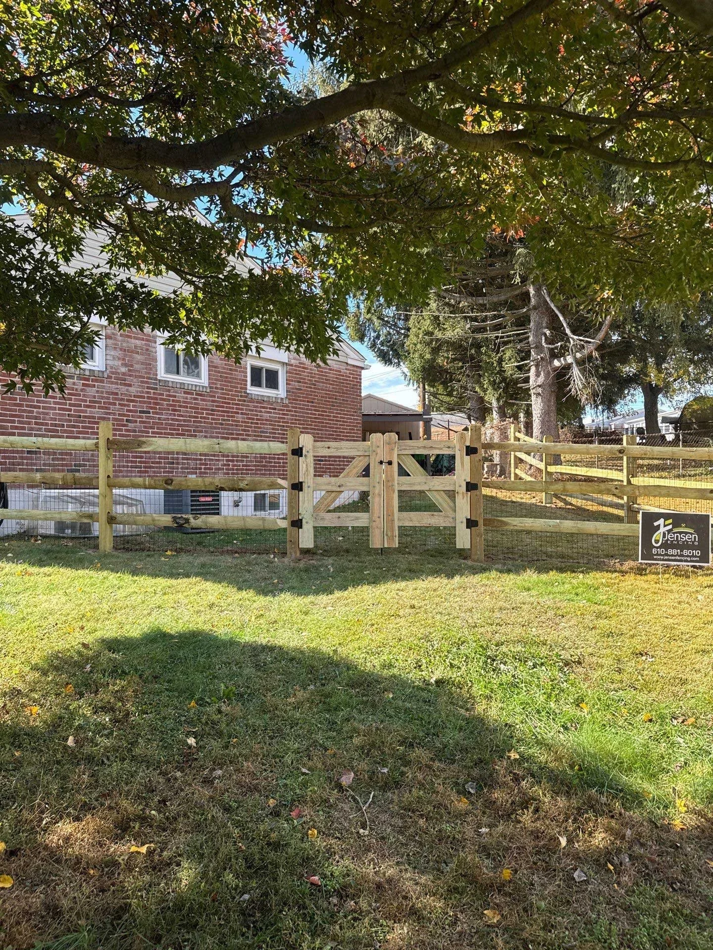 A backyard scene with a wooden fenced yard, a gate in the center, a large tree with green leaves overhead, and a brick house in the background. There is a sign on the fence that reads 'Jensen' with a phone number and website.