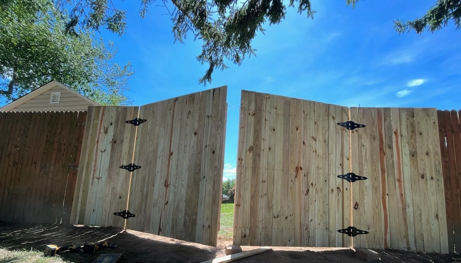A newly constructed wooden fence with steel hinges and an open gate, under a bright blue sky with some trees and a house in the background.