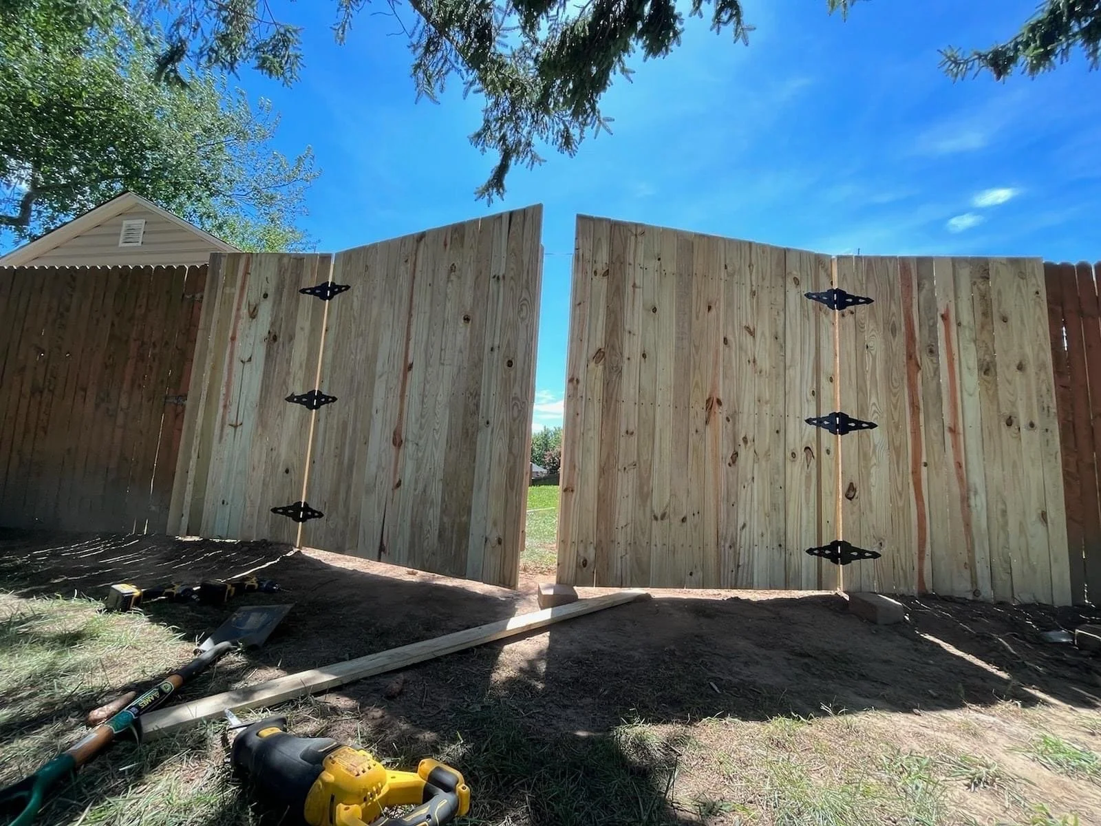 Wooden fence under construction with two panels installed, tools lying on the ground, and a house in the background.