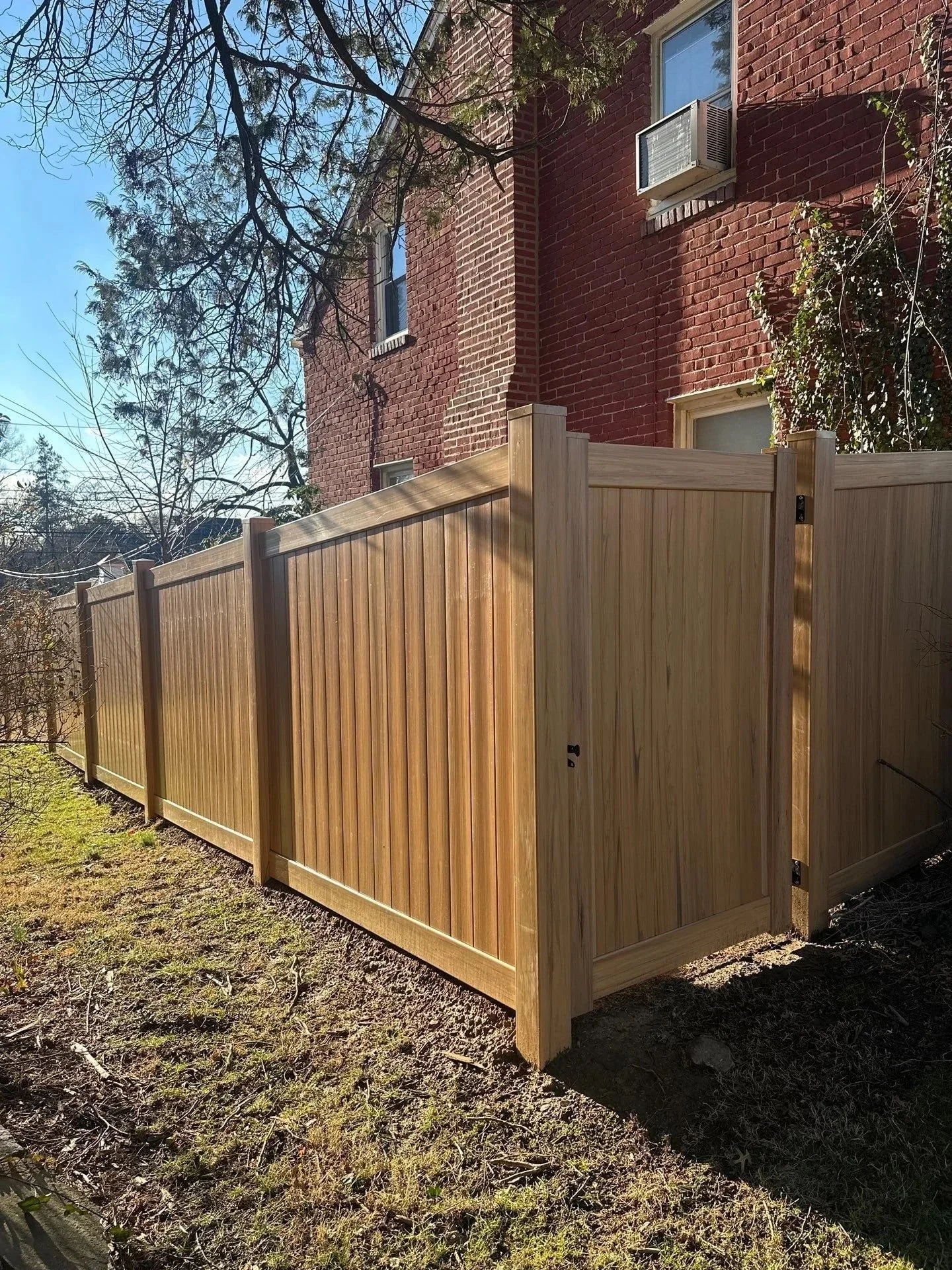 New wooden privacy fence along a backyard next to a red brick building with windows and air conditioning units, under a clear blue sky with leafless trees.