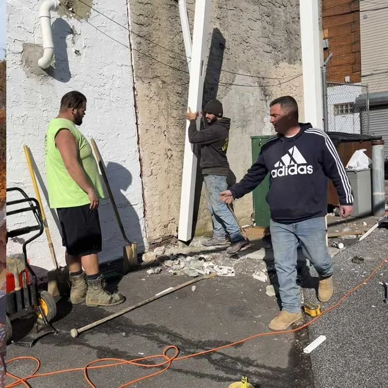 Three men working outside on a construction or demolition project. One man in a black Adidas hoodie is holding a tool and standing on the sidewalk. Another man is carrying a large white panel, while the third man stands nearby with a shovel. Construction debris and tools are scattered on the ground.