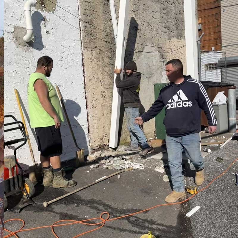 Three men working outside near a wall, with tools and construction debris on the ground. One man is holding a shovel, another appears to be leaning against a large piece of wood or material, and the third is walking toward them.