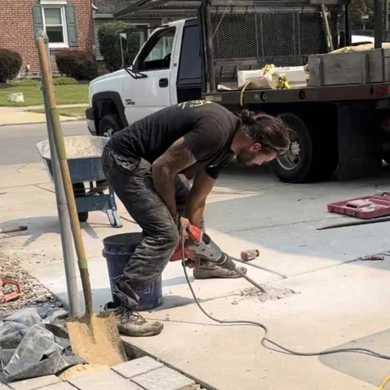 A construction worker wearing black clothing and safety glasses is bending over while using a power saw to cut a concrete sidewalk.