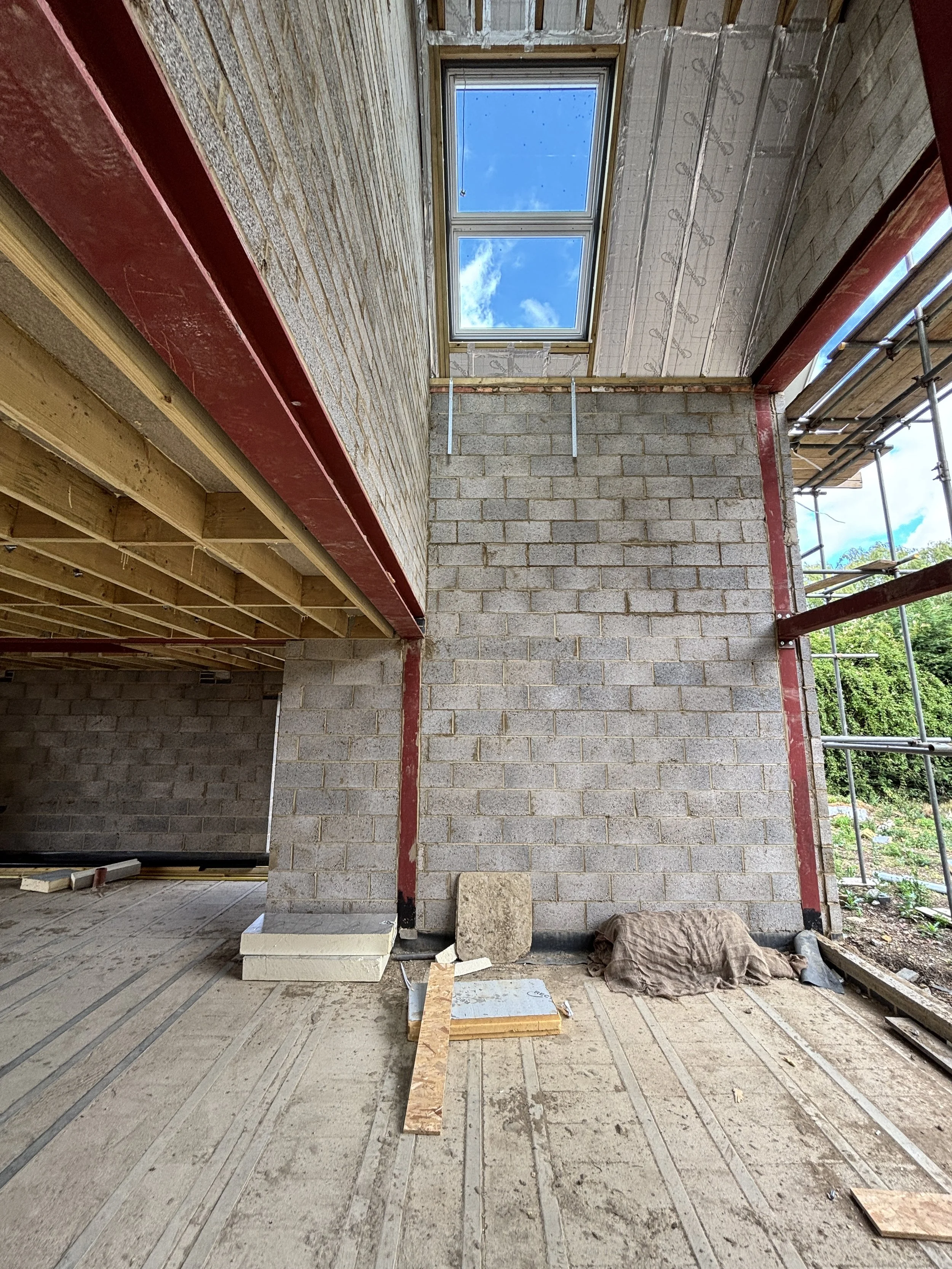 Interior view of a building under construction, showing exposed brick walls stained with red markings, a window, scaffolding outside, wooden framing, and construction materials and debris on the floor.