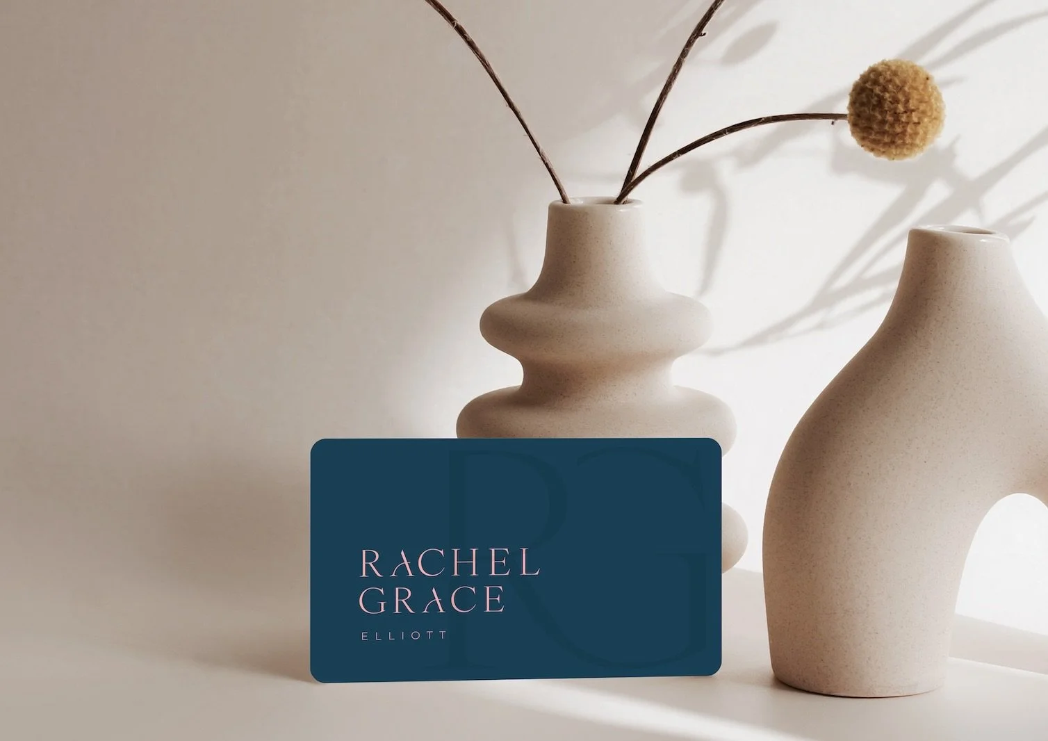 A blue business card with pink text reads 'Rachel Grace Elliott' placed in front of two beige vases with the larger vase containing dried plants all against a neutral background. Coaching branding and design by Hubble.