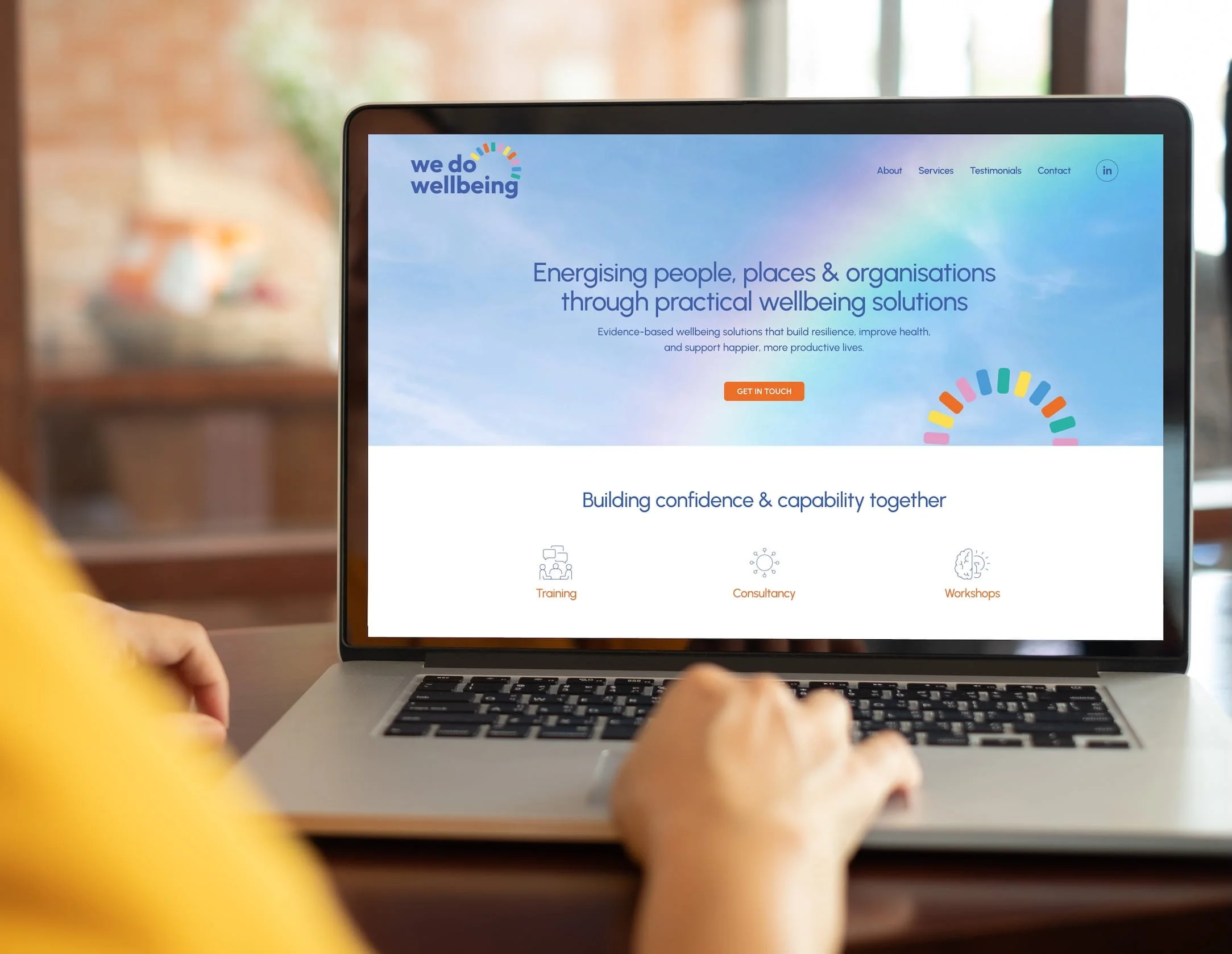 Close-up of a person’s hands typing on a laptop keyboard, displaying a website for 'we do wellbeing' with a blue sky background and colorful logo. Design by Hubble.