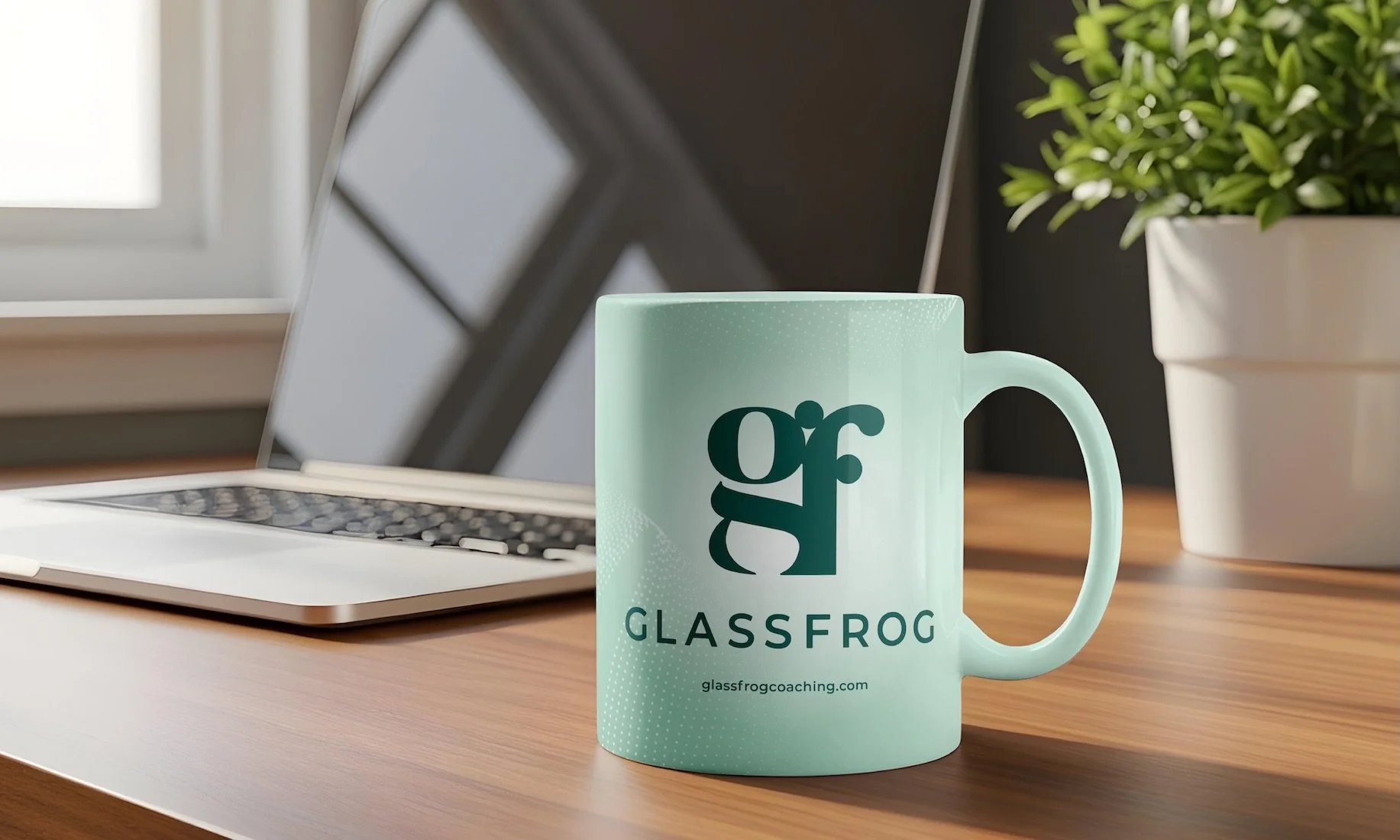 A mint green coffee mug with the GlassFrog logo and website, placed on a wooden desk next to a laptop and a white potted plant in bright indoor lighting. Design by Hubble.