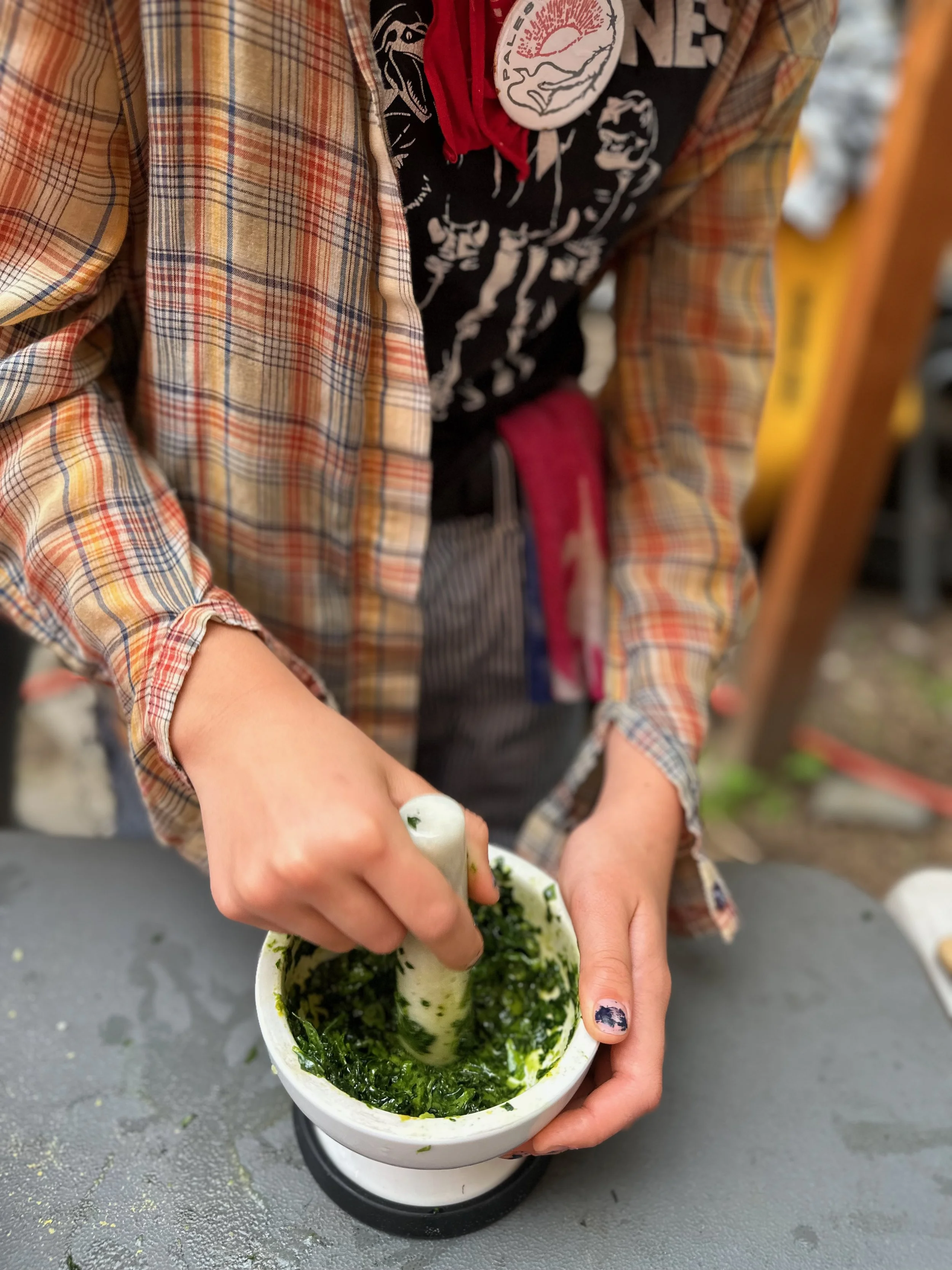 A person in a plaid shirt and black T-shirt with patches is grinding green herbs in a white mortar with a pestle. The person has painted fingernails, and there is a gray surface underneath.