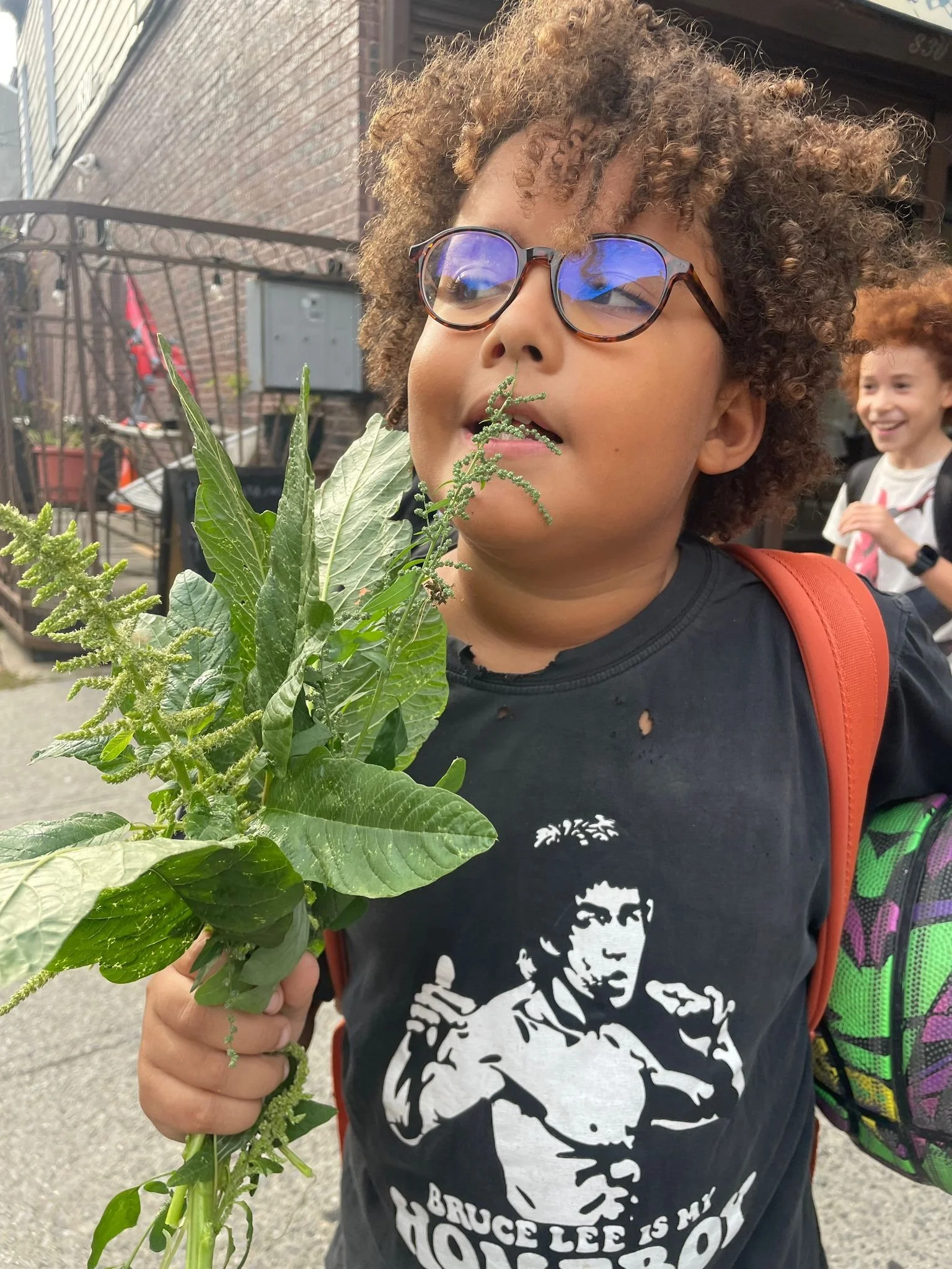 A young boy with curly hair, glasses, and a Bruce Lee t-shirt, holding a bunch of green plants, standing outdoors with a smiling girl in the background.