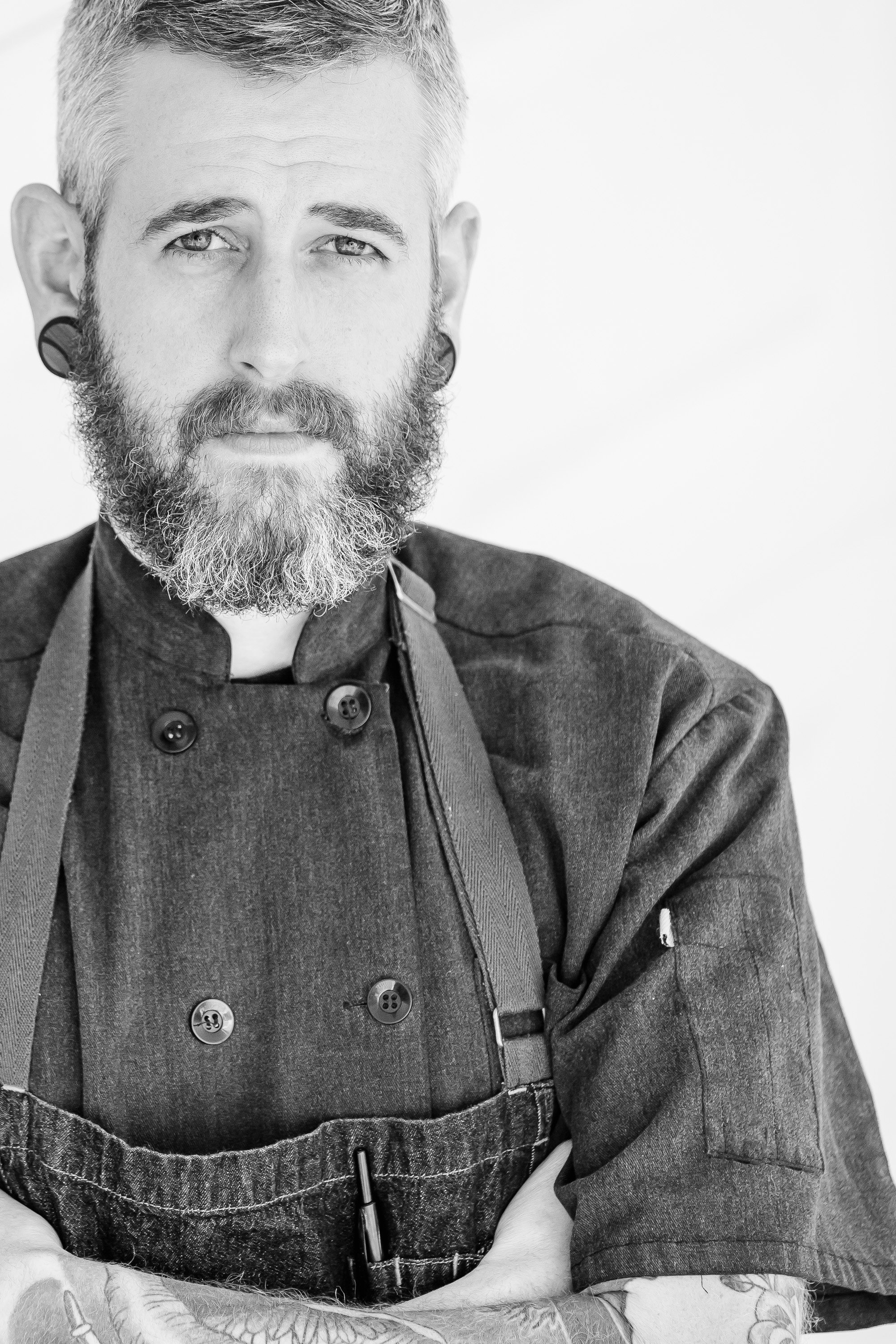Bearded man in a chef's coat and apron, arms crossed, black and white photo.