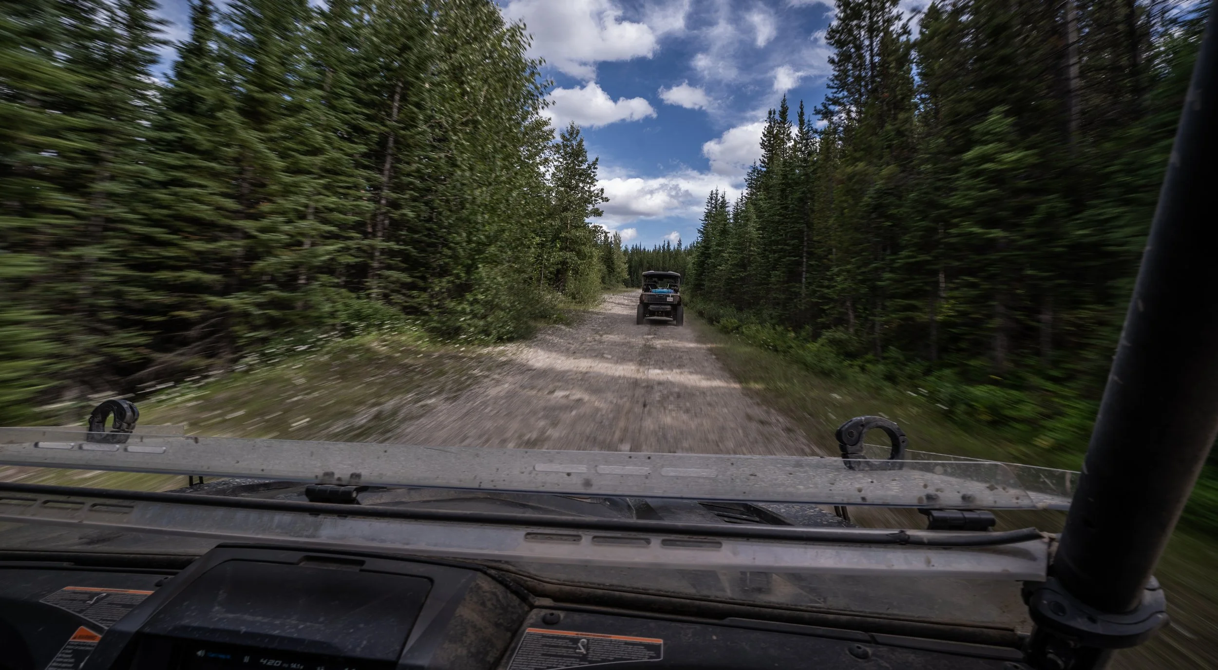View from inside a Yamaha off-road vehicle driving on a dirt trail in a forest with two other Yamaha off-road vehicles ahead, surrounded by tall pine trees and partly cloudy sky.