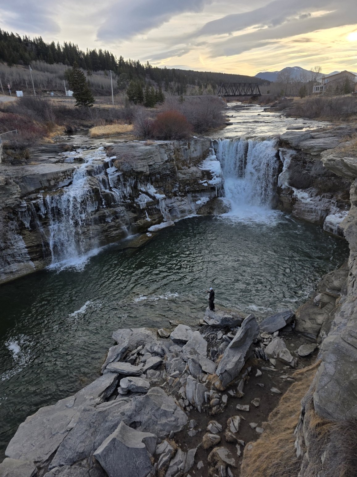 A person standing on rocks in front of a waterfall with cascading water and a pool, surrounded by rocky terrain and trees, under a cloudy sky during sunset.