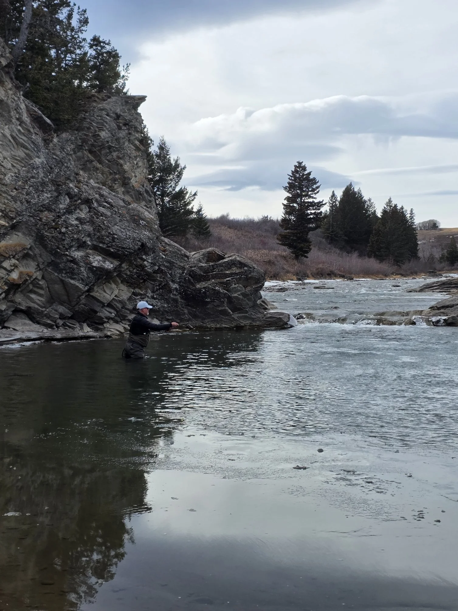 A person wearing a white cap and dark clothing fishing in a river surrounded by rocky cliffs and trees with a cloudy sky overhead.