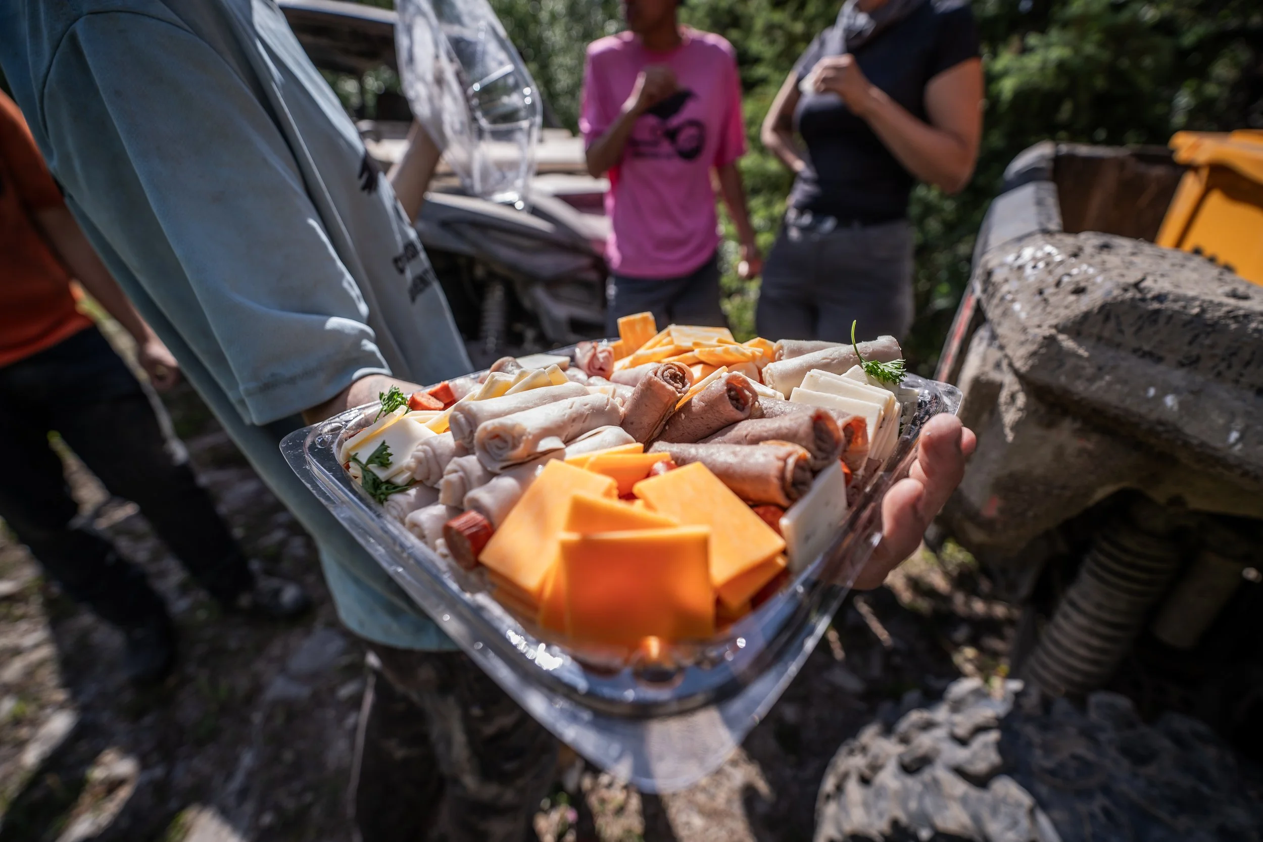 A person holding a tray of cheese, rolled meats, and garnishes outdoors, with other people in the background and a motorcycle nearby.