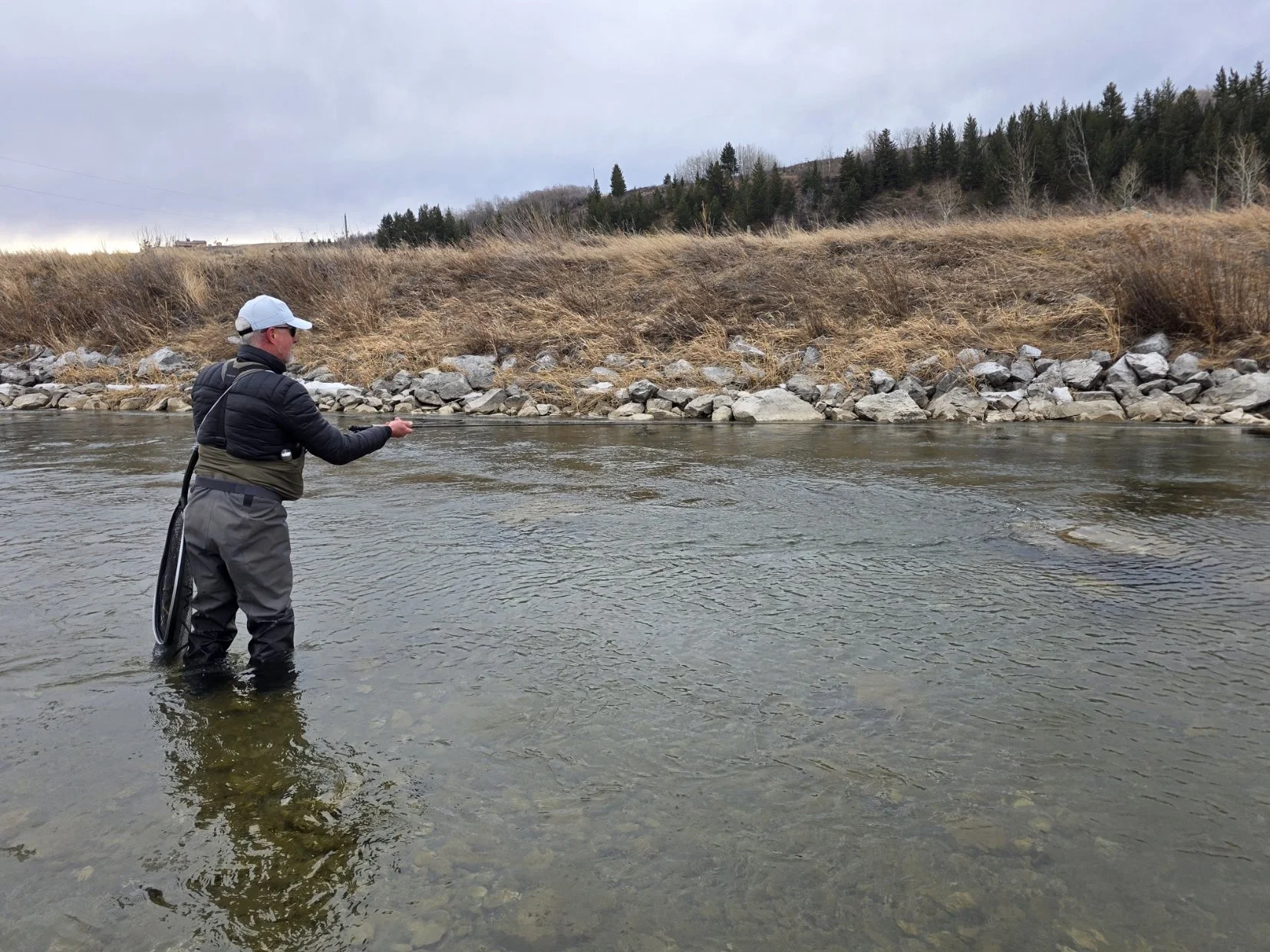 A person fishing in a river, wearing waterproof waders, a puffy jacket, and a white cap, with a forested hillside and cloudy sky in the background.